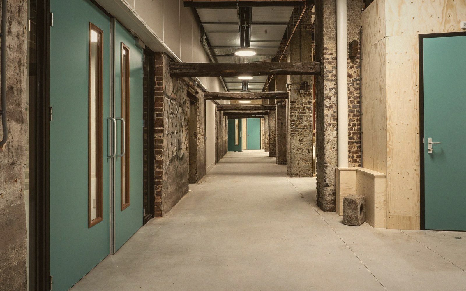 Exposed brick columns and timber beams line a corridor with teal doors and concrete flooring